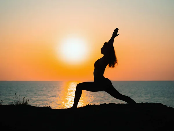 Молода струнка дівчина практикує йогу на горі проти океану або моря під час сходу сонця. Силует жінки в променях чудового заходу сонця . — стокове фото
