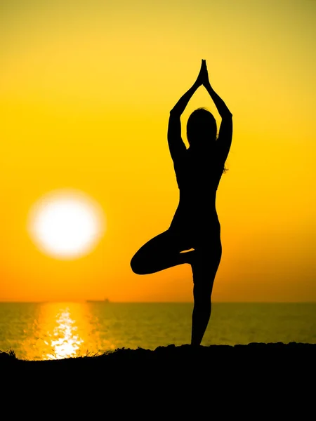 Young slim girl practicing yoga on mountain against ocean or sea at sunrise time. Silhouette of woman in yellow rays of awesome sunset. — Stock Photo, Image