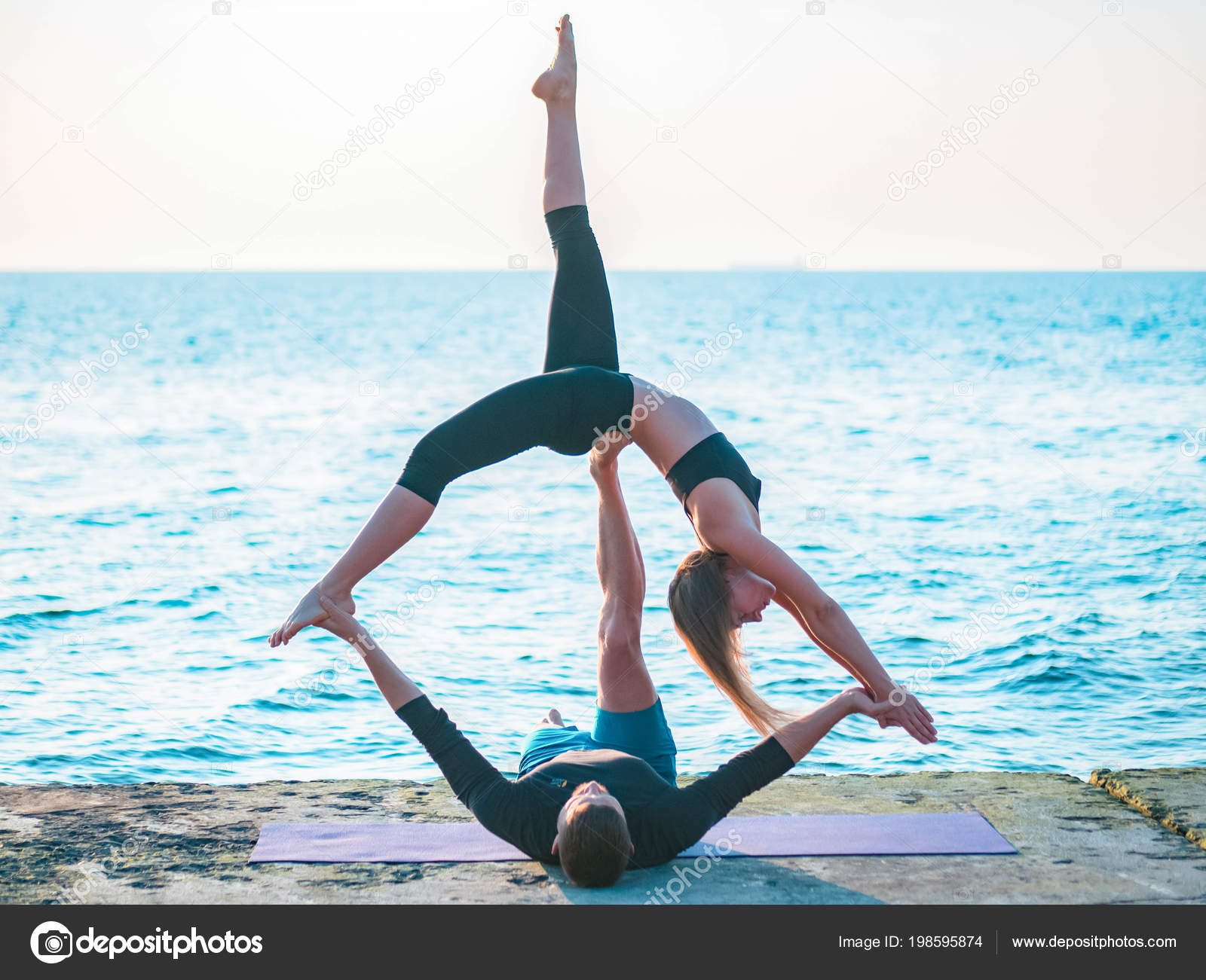 Fit jovem casal fazendo acro-ioga na praia do mar. Homem deitado em placas  de cimento e mulher equilibrada em seus pés. Belo par praticando ioga  juntos . — Foto © kohanova1991 #198595874, image size:1600x1301