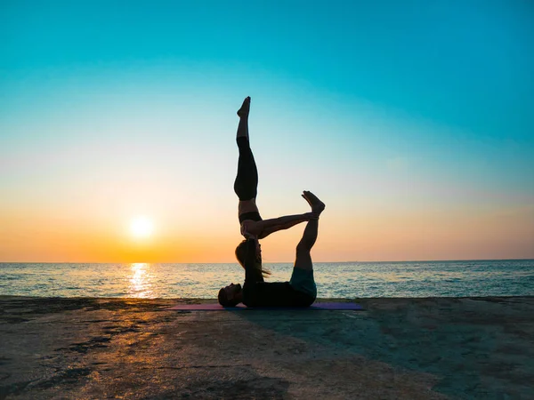 Silhouettes de jeunes couples en forme faisant de l'acro-yoga à la plage de la mer. Un homme allongé sur des plaques de béton et une femme équilibrée dans ses pieds. Beau couple pratiquant le yoga ensemble — Photo