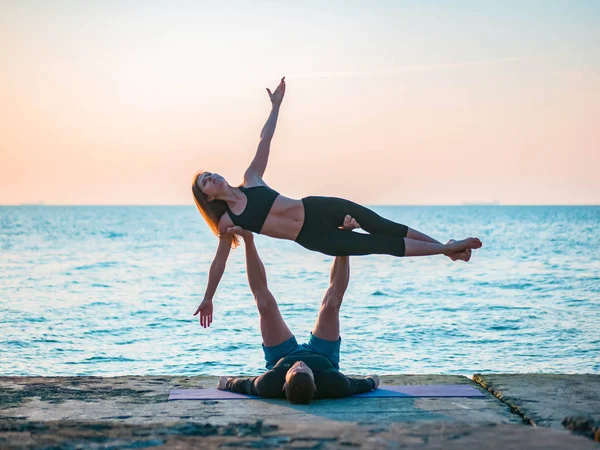 Adatto giovane coppia che fa acro-yoga sulla spiaggia di mare. Uomo sdraiato su piastre di cemento e bilanciamento donna in piedi. Bella coppia praticare yoga insieme . — Foto Stock