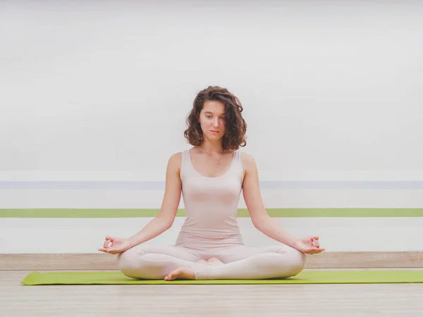 Joven mujer bonita practicando yoga en la sala de clase de estudio minimalista de luz. Chica atractiva se sienta en posición de loto clásico y comienza su meditación — Foto de Stock