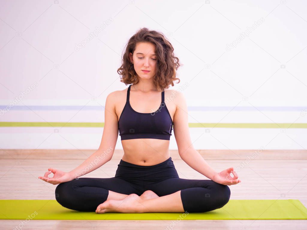 Young pretty woman practicing in yoga at light minimalist studio class room. Attractive girl sits in classic lotus position and begins her meditation