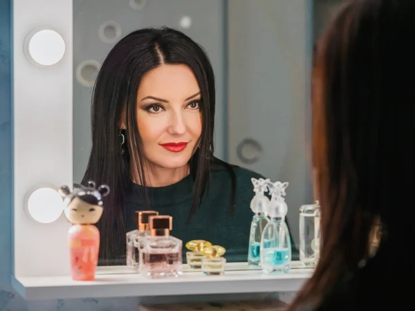 Portrait of beautiful, mature and sensual woman with elegant hairstyle looking at her reflection in mirror and fixing hair. Makeup at night getting ready before going to party. — Stock Photo, Image