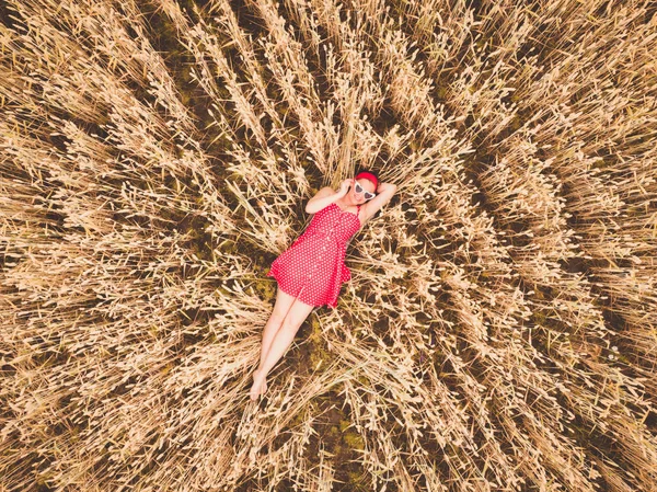 Young beautiful woman in red retro dress and sunglasses lying in wheat yellow field. Flying close above cornfield. AERIAL Drone view. Harvest, agriculture concept.