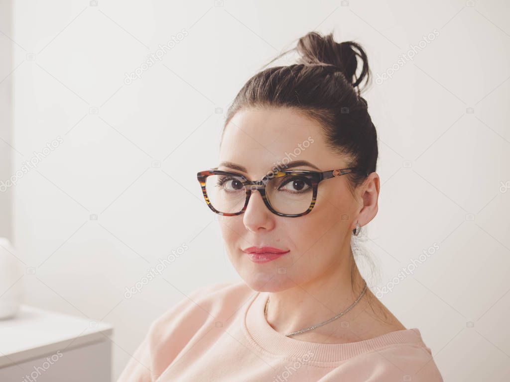 Beautiful mature woman in big glasses smiles and looks to camera. Face portrait of adult lady at home.