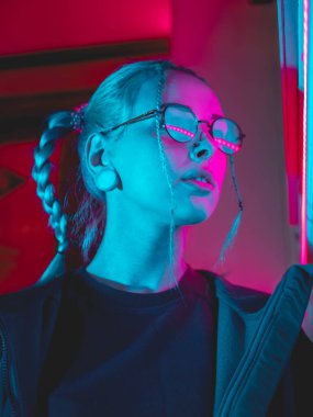 Şehrin gece parlayan pembe ve mavi neon ışıkları yakınında alışılmadık saç modeli genç güzel kız. Mavi boyalı saç örgü içinde. Ciddi hipster genç gözlük ve güzel lensler.