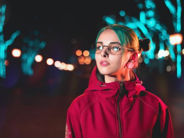 Neon ışıkları arka plan ile sokak geceleri ayakta piercing genç güzel saçlı kız mavi boyalı alışılmadık, şeffaf gözlük. Mutlu sevimli şık genç portresi.