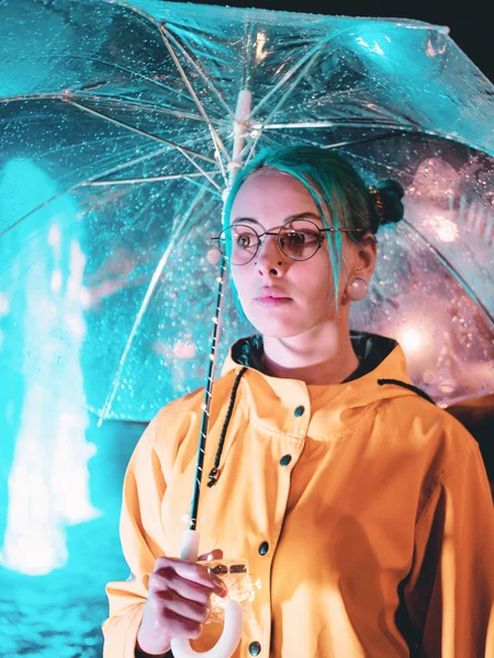 Genç güzel kız sarı yağmurluk mavi boyalı saçlı ve şeffaf şemsiye ile Çeşme duruyor. Şehrin gece neon aydınlatma. Gözlüklü şık hipster portresi. — Stok fotoğraf