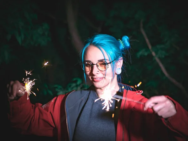Potret gadis hipster dengan rambut berwarna biru dengan kilauan di tangan dan refleksi dalam kacamata. Remaja muda yang cantik berdiri di kota malam, bersenang-senang dengan api bengal . — Stok Foto