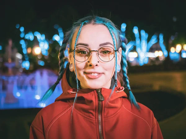 Krásná mladá dívka s modrou obarvená neobvyklé, průhledné brýle, piercing stojící v noci ulici s neony pozadím. Portrét šťastný roztomilý elegantní teenager. — Stock fotografie