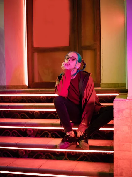 Genç güzel kız merdivende hangi parlak kırmızı otururken sıradışı saç modeli ile neon ışıkları. Gece şehir. Mavi boyalı saç örgü içinde. Mutlu hipster genç gözlük.