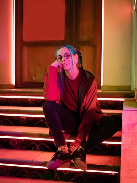 Genç güzel kız merdivende hangi parlak kırmızı otururken sıradışı saç modeli ile neon ışıkları. Gece şehir. Mavi boyalı saç örgü içinde. Mutlu hipster genç gözlük.