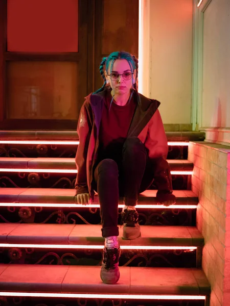 Genç güzel kız merdivende hangi parlak kırmızı otururken sıradışı saç modeli ile neon ışıkları. Gece şehir. Mavi boyalı saç örgü içinde. Mutlu hipster genç gözlük.