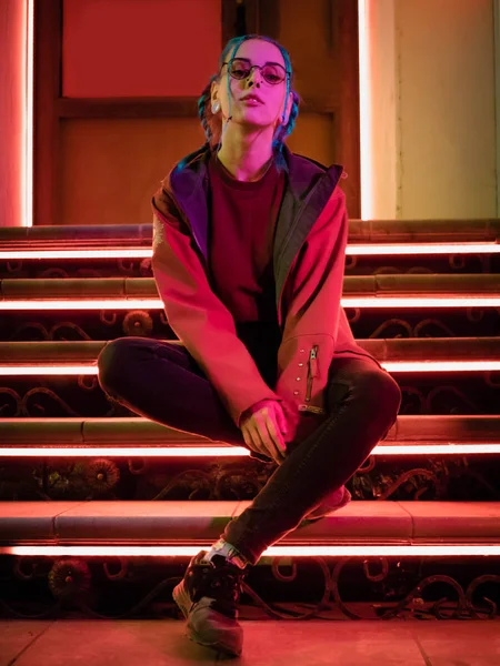 Menina bonita nova com penteado incomum sentado em escadas que brilham luzes de néon vermelho. Cidade à noite. Cabelo azul tingido em tranças. adolescente hipster feliz em óculos . — Fotografia de Stock