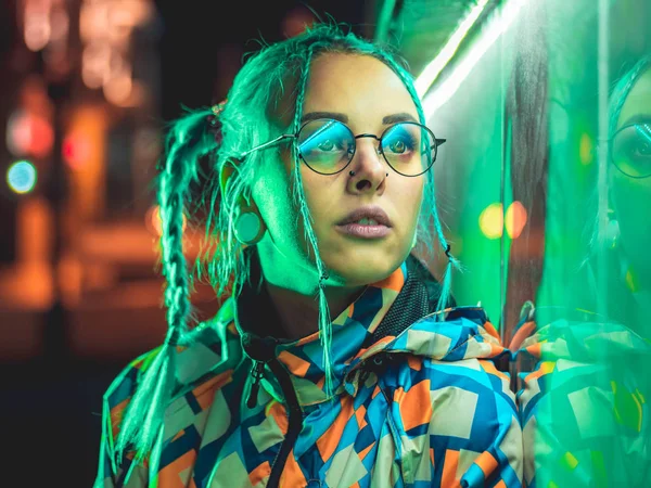 Νέοι όμορφο κορίτσι με ασυνήθιστη χτένισμα κοντά σε λαμπερό πράσινο φως νέον της πόλης τη νύχτα. Βαμμένα μπλε μαλλιά σε πλεξούδες. Συλλογισμένος hipster Έφηβος με τα γυαλιά. — Φωτογραφία Αρχείου