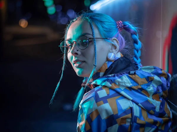 Νέοι όμορφο κορίτσι με ασυνήθιστη χτένισμα κοντά στο λαμπερό μπλε φως νέον της πόλης τη νύχτα. Βαμμένα μπλε μαλλιά σε πλεξούδες. Συλλογισμένος hipster Έφηβος με τα γυαλιά. — Φωτογραφία Αρχείου