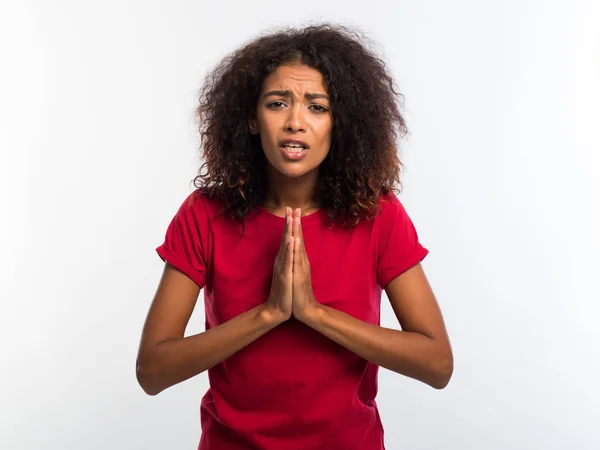 Приваблива африканська жінка в червоній футболці молиться і схрещує пальці на білому тлі. Дівчина змішаної раси благає когось . — стокове фото