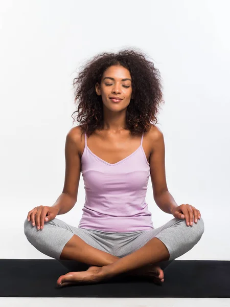 Mujer afroamericana feliz en deportes cómodos llevar meditando en la estera sobre fondo blanco de la pared. Chica negra concentrada en la práctica de yoga . — Foto de Stock