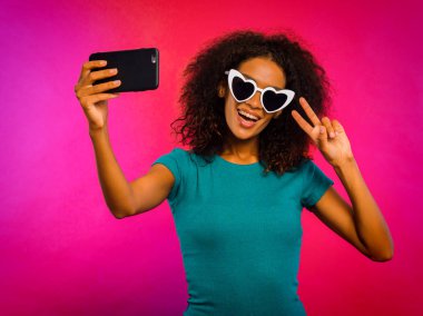 Mutlu genç Afro-Amerikan Kız kalbinde güneş gözlüğü ve yeşil t-shirt yapımı selfie resmi smartphone için pembe parlak arka plan üzerinde şekillendiriyor. Teknoloji kavramı.