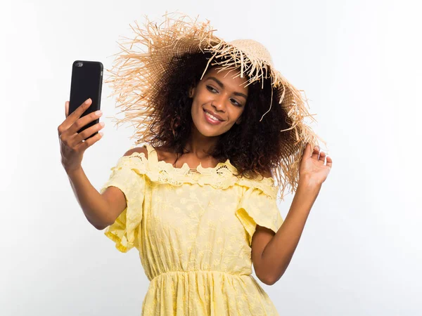 Lächelnd glückliche junge afrikanisch-amerikanische Mädchen in gelbem Kleid und Strohhut machen Selfie-Foto auf Smartphone über weißem Hintergrund. Technologiekonzept. — Stockfoto