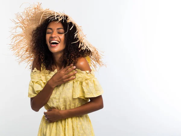 행복 한 흑인 여자 곱슬 헤어스타일 머리 노란 드레스와 밀 짚 모자와 흰 벽 배경 위에 contagiously 웃는 다. 여자 재미, 유혹, 카메라에 웃 고. — 스톡 사진