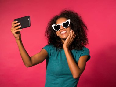 Gülümseyen mutlu genç Afro-Amerikan kız yeşil t-shirt ve kalp selfie fotoğraf smartphone üzerinde pembe arka plan yapma güneş gözlüğü şeklinde. Teknoloji kavramı.