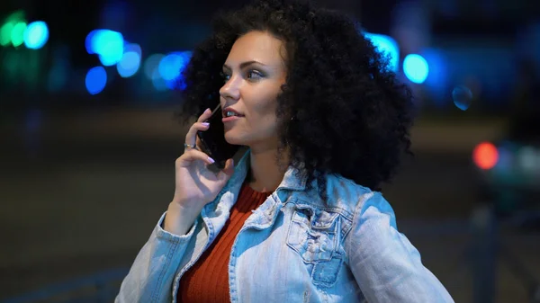 Young beautiful woman with very curly afro hair talking on her mobile phone at night illuminated street. Trendy girl have conversation. Smartphone, technology concept.