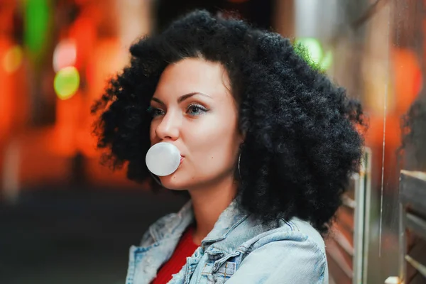 Çok kıvırcık afro saçlı Genç güzel kadın bir kabarcık top beyaz sakız geceleri açılırsa. Trendy kız eğleniyor, hayattan zevk. Güzel ışıklı sokak.