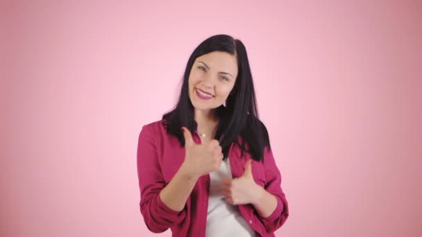 Vencedor. Sucesso. Menina morena em camisa rosa no fundo claro sorri para a câmera e dá polegares para cima. Mulher feliz dançando em câmera lenta — Vídeo de Stock