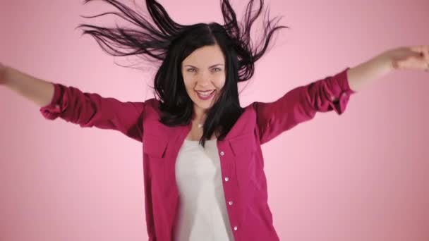 Chica de la energía sobre fondo rosa en el estudio. Lleva una camisa colorida. Cabello negro en la cola está volando desde el movimiento. Movimiento lento — Vídeos de Stock