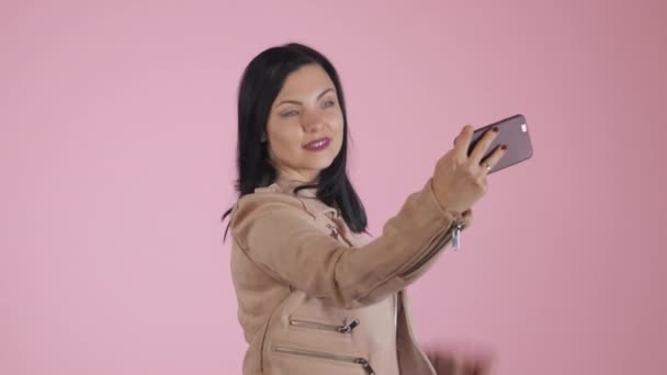 Sonriente mujer morena feliz en chaqueta rosa haciendo selfie en el teléfono inteligente sobre fondo colorido . — Vídeo de stock