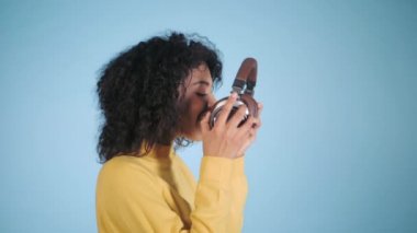 Afrikalı kadın mavi renkli arka plan üzerinde müzik dinlemek. Kız sarı giyim üzerinde kulaklık koymak ve melodiler tadını çıkarırken. Ağır çekim