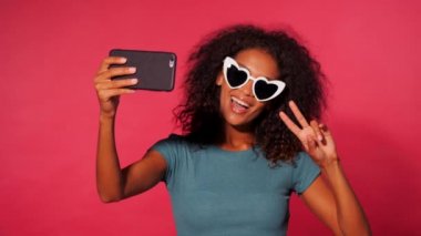 Pembe duvar arka plan üzerinde gülümseyen mutlu Afrikalı-Amerikalı kadın Smartphone cep telefonu ile yeşil t-shirt yapımı selfie kıvırcık saçlı. Kalp şeklinde güneş gözlüklü kız.