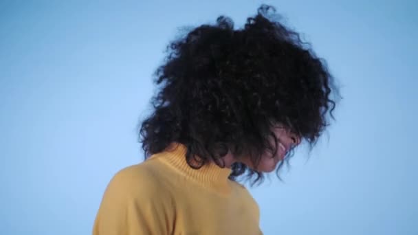 Mooie zwarte vrouw met afro haar plezier glimlachend en dansen in de studio tegen blauwe achtergrond. Slow motion. — Stockvideo