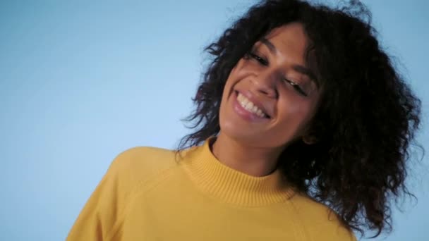 Beleza sorrindo mulher de raça mista em roupas amarelas olhando para a câmera, toca seu cabelo afro e dançando em estúdio contra fundo azul. câmara lenta . — Vídeo de Stock