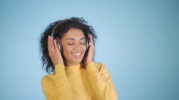 Słuchanie muzyki na niebieskim tle kolorowe kobieta mulat. Mix wyścig dziewczyna z afro włosów kręconych w studio, zwolnionym tempie. — Wideo stockowe
