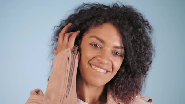 Jovem mulher brasileira africana retrato posando sobre fundo azul claro. Estilo moderno e urbano, jaqueta de moda rosa, penteado afro. Ela morde os lábios e parece sexy. Movimento lento . — Vídeo de Stock