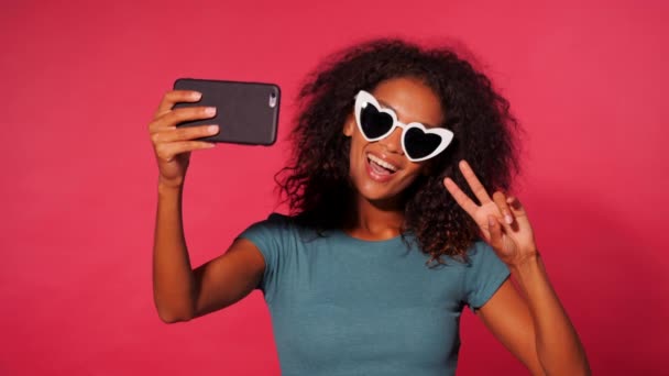 Lächelnde fröhliche afrikanisch-amerikanische Frau mit lockigem Haar in grünem T-Shirt, die vor rosa Wandhintergrund ein Selfie mit dem Smartphone macht. Mädchen mit herzförmiger Sonnenbrille. — Stockvideo