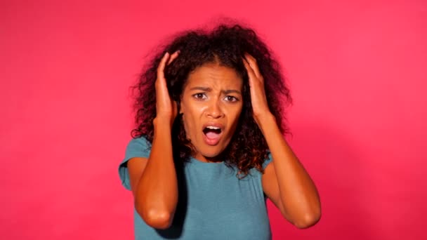 Frightened african american woman with curly hair in green t-shirt afraid of something and looks into the camera with big eyes full of horror over pink wall background. — Stock Video