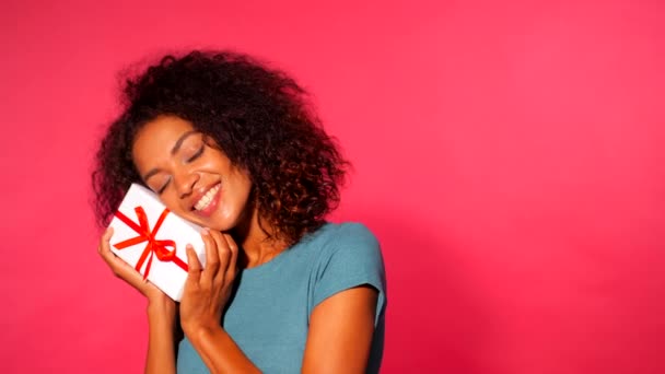 Jolie jeune femme afro-américaine tenant boîte cadeau avec ruban rouge et arc sur fond mural rose. Fille souriante, elle est heureuse d'être présente . — Video