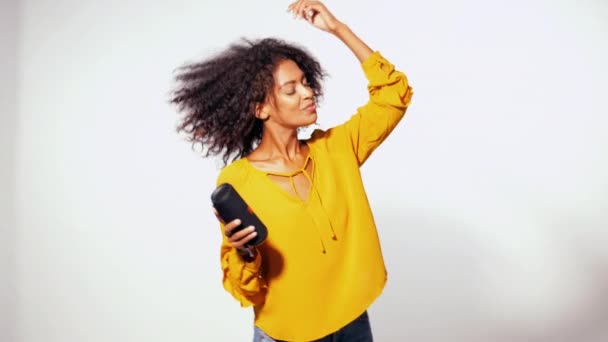 Young beautiful african american woman enjoying and dancing at white background. Modern trendy girl with afro hairstyle listening to music by wireless portable speaker. — Stock Video