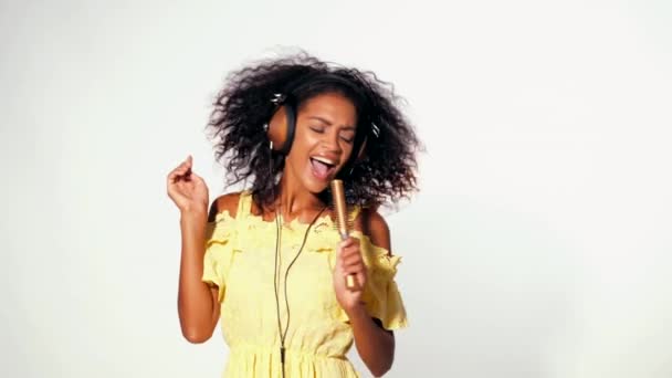 Jeune femme afro-américaine mignonne en robe jaune chantant et dansant avec sèche-cheveux à la place microphone à fond blanc. Moderne fille à la mode avec coiffure afro . — Video
