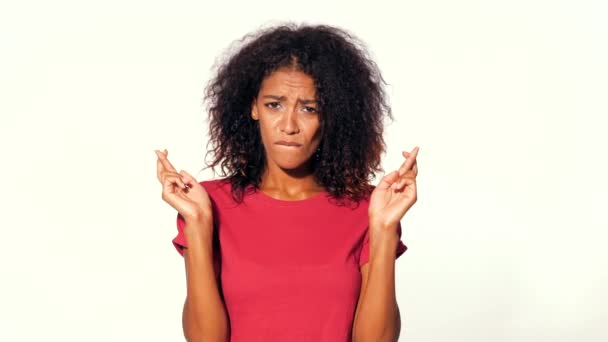 Atractiva mujer africana en camiseta roja rezando y cruza sus dedos sobre fondo blanco. Chica de raza mixta rogando a alguien . — Vídeos de Stock