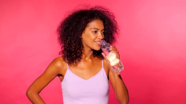 Attractive african-american sporty young woman drinking bottle of water on pink wall background. Girl in sports clothes after fitness. Studio footage. Slow motion. — Stock Video