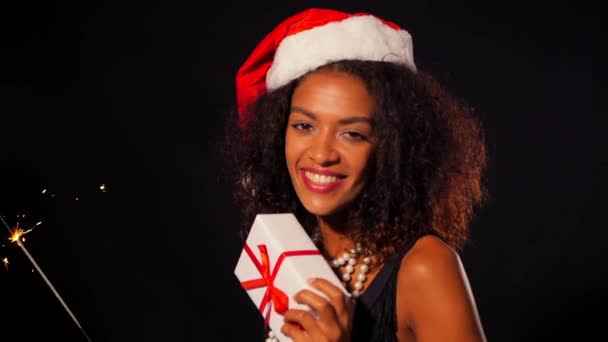 Afro-americana giovane donna in abito da festa in possesso di scatola regalo e bengala scintille fuoco su sfondo luci nere. Ragazza in cappello di Babbo Natale, felice di ricevere un regalo a Capodanno o Natale . — Video Stock