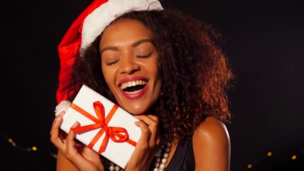 Afrikaans-Amerikaanse jonge vrouw in partij jurk en KERSTMUTS holding geschenkdoos met rood lint op zwarte achtergrond. Meisje glimlachend, ze krijgen graag presenteren op Nieuwjaar of Christmas. — Stockvideo