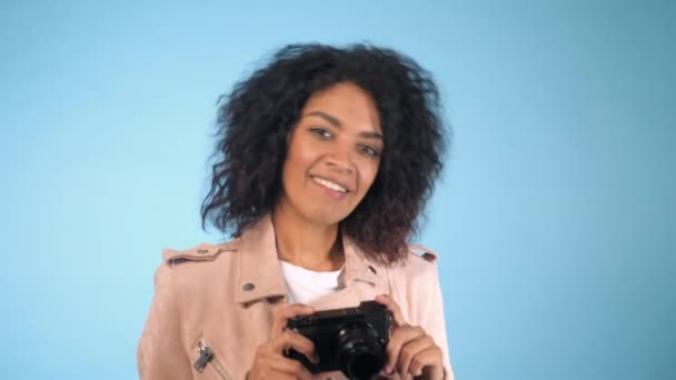 Joven mujer africana bonita en chaqueta rosa toma fotos con cámara réflex digital sobre fondo azul en el estudio. Chica sonriendo, coqueteando y divirtiéndose como fotógrafo . — Vídeos de Stock