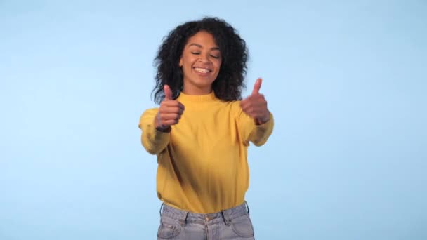 Winner. Success. Positive brunette young african girl on light blue background smiles for camera and gives thumbs up. Happy woman dancing — Stock Video