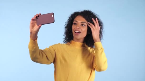 Красивая африканская американка держит и использует смартфон, чтобы снять себя в синей студии. Чернокожая женщина использует технологию сетевого общения на отдыхе, образ жизни . — стоковое видео